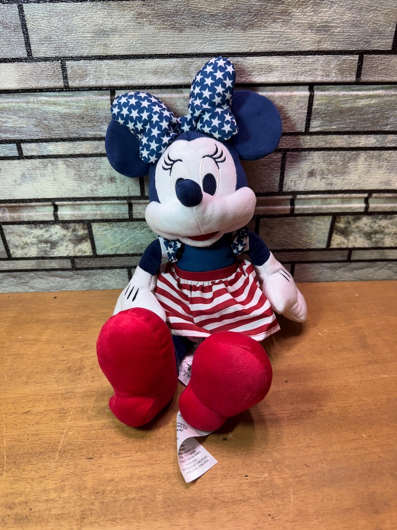 May include: A Minnie Mouse plush doll with a blue bow featuring white stars, blue ears, and a red and white striped skirt. The doll has red shoes and white gloves. The doll is sitting on a wooden surface.
