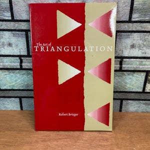 Op de afbeelding: Een hardcover boek getiteld "The Art of Triangulation" door Robert Brieger. De omslag heeft een rood en beige ontwerp met witte en roze driehoeken. Het boek ligt op een houten oppervlak.