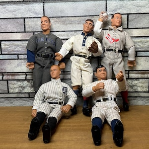 May include: Five baseball player action figures wearing vintage baseball uniforms. The figures are posed in a variety of stances, including batting, pitching, and fielding. The figures are made of plastic and have detailed features, including realistic facial expressions and clothing.