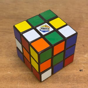 Könnte beinhalten: Ein mehrfarbiger Rubik's Cube mit grünen, gelben, orangefarbenen, weißen, blauen und roten Quadraten. Der Würfel hat ein zentrales Logo. Ein klassisches Puzzlespielzeug.