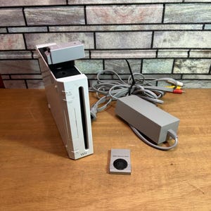 Op de afbeelding: Een witte Nintendo Wii-console met een voedingsadapter, kabels en een geheugenkaart. De console staat rechtop, met het Wii-logo zichtbaar. De voedingsadapter en kabels zijn grijs. De geheugenkaart is zilverkleurig.