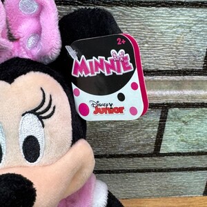 Puede incluir: Un peluche de Minnie Mouse con lazo rosa, vestido rosa y detalles negros. La etiqueta dice "Minnie" y "Disney Junior". Dise&ntilde;ado para ni&ntilde;os de 2 a&ntilde;os en adelante.