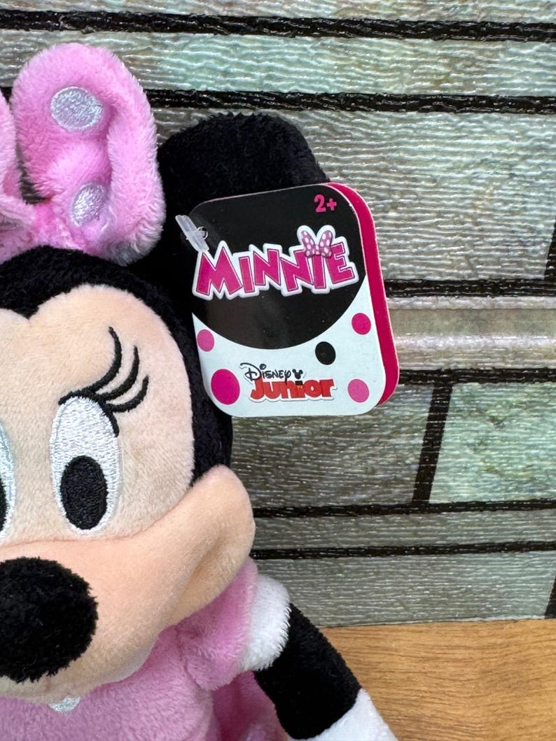 Puede incluir: Un peluche de Minnie Mouse con lazo rosa, vestido rosa y detalles negros. La etiqueta dice "Minnie" y "Disney Junior". Dise&ntilde;ado para ni&ntilde;os de 2 a&ntilde;os en adelante.
