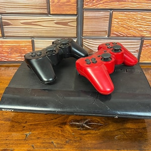 May include: A black Sony PlayStation 3 game console with a ribbed top surface and a glossy front panel featuring the Sony logo. Two wireless game controllers, one in solid black and one in solid red, are resting on top of the console unit.