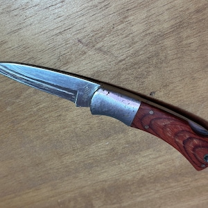 May include: A small pocket knife with a wood handle and a silver blade. The blade has a Damascus pattern and the handle has a dark wood grain.