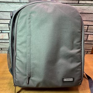 May include: A gray backpack with a top handle, side mesh pocket, and a front zipper. The backpack has a small pocket on the front and a "CADEN" label. The bag is sitting on a wooden surface.