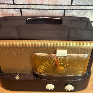 K&ouml;nnte beinhalten: Ein vintage AM/FM-Radio in Schwarz und Gold mit einem runden Zifferblatt und zwei Kn&ouml;pfen. Das Radio hat ein Lautsprechergitter an der Vorderseite und eine kleine Metallplatte mit einem Logo an der Unterseite.