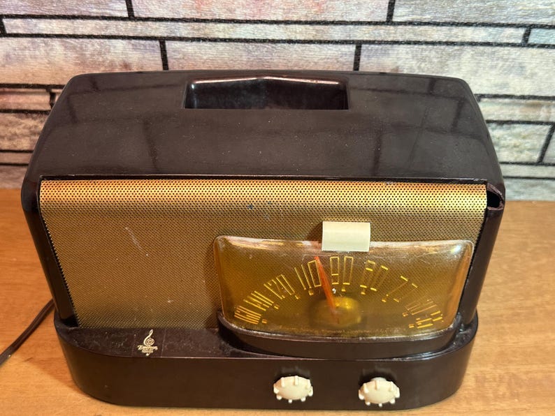 K&ouml;nnte beinhalten: Ein vintage AM/FM-Radio in Schwarz und Gold mit einem runden Zifferblatt und zwei Kn&ouml;pfen. Das Radio hat ein Lautsprechergitter an der Vorderseite und eine kleine Metallplatte mit einem Logo an der Unterseite.
