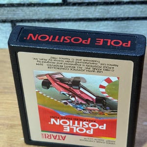 Puede incluir: Un cartucho de videojuego Atari negro con una etiqueta roja y blanca. La etiqueta presenta un coche de carreras rojo en una pista con el texto "POLE POSITION" en letras blancas grandes. El texto "FAST-PACED TRACK FUN!" también está en la etiqueta.