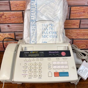 Puede incluir: Un fax Brother MFC 970MC de color beige con auricular telefónico, teclado numérico y varios botones de función. El dispositivo incluye una pantalla digital y viene acompañado de un manual de usuario en una funda de plástico y un adaptador.