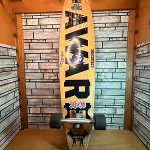 Könnte beinhalten: Ein Longboard-Skateboard mit schwarzen Rollen und einem gelben Deck. Das Deck hat eine schwarz-weiße Grafik mit dem Wort "Aware" mit einer Ziege und den Texten "Big/DC" und "Iron Maiden". Auf dem Deck befinden sich Aufkleber für "Sex Pistols" und "Iron Maiden".