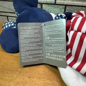 May include: A plush toy with a blue head and body, featuring white stars on blue fabric. The toy has red and white striped fabric on the side. A tag with multilingual warnings is attached.