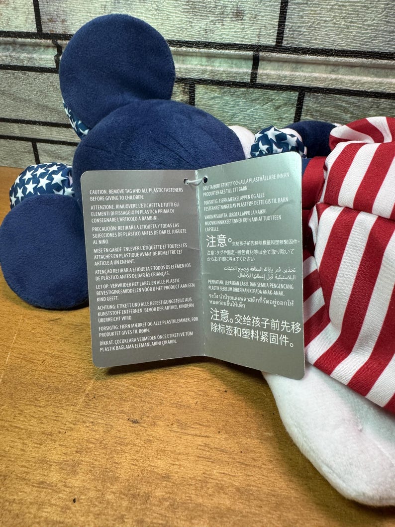 May include: A plush toy with a blue head and body, featuring white stars on blue fabric. The toy has red and white striped fabric on the side. A tag with multilingual warnings is attached.
