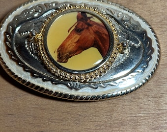 May include: A silver and gold belt buckle with a white enamel background and a detailed design. The buckle features a round image of a brown horse's head.