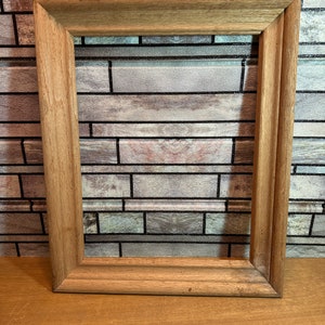 May include: A rectangular wooden picture frame with a natural wood finish. The frame has a simple, classic design with a beveled edge. The frame is empty, ready to hold a photograph or artwork.