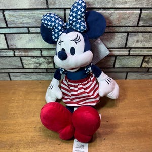 May include: A plush Minnie Mouse doll in a seated position. She wears a blue dress with a red and white striped skirt, a blue bow with white stars, and red shoes. Her white gloves and black eyes are visible.