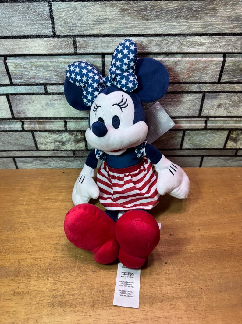 May include: A plush Minnie Mouse doll in a seated position. She wears a blue dress with a red and white striped skirt, a blue bow with white stars, and red shoes. Her white gloves and black eyes are visible.