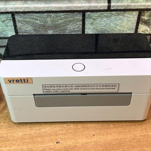 May include: A white and black Vretti thermal label printer with a rectangular body, a glossy black top panel, and a circular power button. A label on the front reads: Please use cotton swab with 100% alcohol to clean the print head and rubber roller.