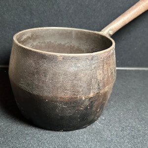 May include: A vintage cast iron pot with a long handle. The pot is dark gray and has a worn, rustic look.