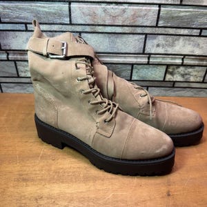 May include: A pair of tan suede ankle boots with a black sole. The boots have a lace-up design, a buckle strap around the ankle, and a small pull tab at the back. The boots are displayed on a wooden surface.