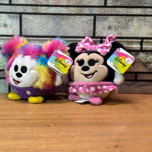 May include: Two Disney plush toys: Mickey Mouse with rainbow fur and Minnie Mouse with a pink polka dot skirt. Both have Disney tags attached. The toys are on a wooden surface with a brick background.