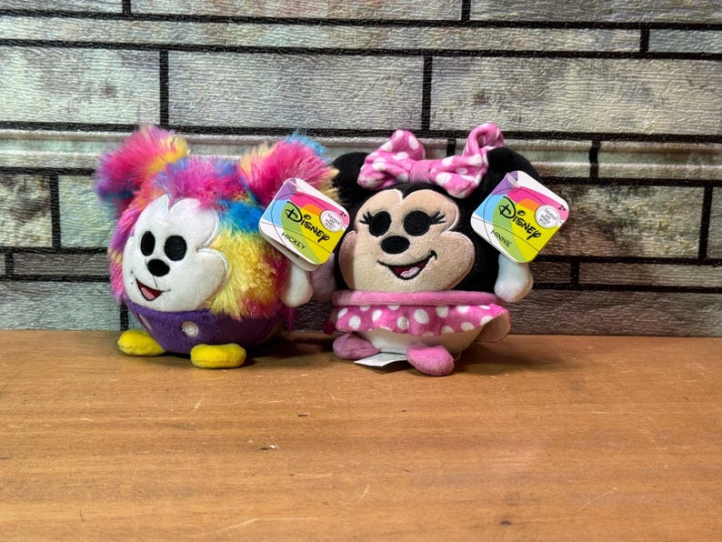 May include: Two Disney plush toys: Mickey Mouse with rainbow fur and Minnie Mouse with a pink polka dot skirt. Both have Disney tags attached. The toys are on a wooden surface with a brick background.