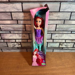Może przedstawiać: Lalka Disney Princess Ariel w fioletowej i zielonej sukience z koroną na głowie. Lalka jest zapakowana w r&oacute;żowe i niebieskie pudełko z logo Disney Princess.