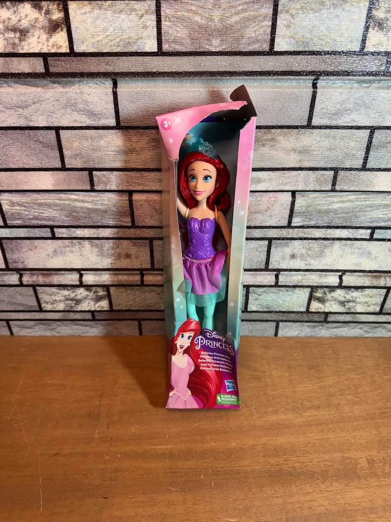 Może przedstawiać: Lalka Disney Princess Ariel w fioletowej i zielonej sukience z koroną na głowie. Lalka jest zapakowana w r&oacute;żowe i niebieskie pudełko z logo Disney Princess.