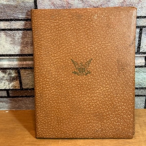 May include: A rectangular book with a textured tan cover featuring a small, centred gold-coloured emblem of an eagle with outstretched wings above a shield. The cover has a pebbled surface texture and the book is positioned vertically.