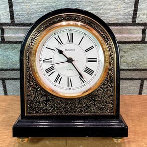 Puede incluir: Un reloj de sobremesa de cuarzo Bulova con marco negro arqueado y esfera de metal dorado con números romanos. El reloj tiene una esfera blanca con manecillas negras y un intrincado detalle de filigrana floral dorada en la base.