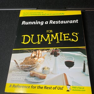 May include: A yellow and black book cover with the title "Running a Restaurant For Dummies". The cover features a photo of a restaurant table setting with bread, butter, and a glass of red wine.