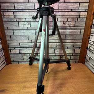 May include: A silver and black Ambico tripod with adjustable legs and a black head. The tripod is designed for cameras and video equipment. The legs are adjustable for height and stability.