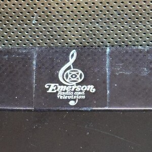 K&ouml;nnte beinhalten: Nahaufnahme eines Vintage-Logos von Emerson Radio and Television mit einem Violinschl&uuml;ssel und einem Kreis mit einem Kreuz darin. Das Logo ist in Silber auf einer dunkelbraunen Oberfl&auml;che gedruckt.