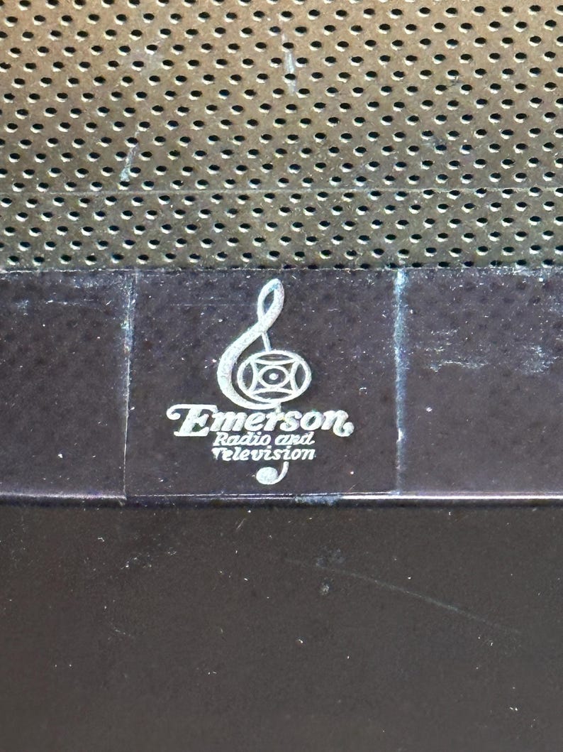 K&ouml;nnte beinhalten: Nahaufnahme eines Vintage-Logos von Emerson Radio and Television mit einem Violinschl&uuml;ssel und einem Kreis mit einem Kreuz darin. Das Logo ist in Silber auf einer dunkelbraunen Oberfl&auml;che gedruckt.