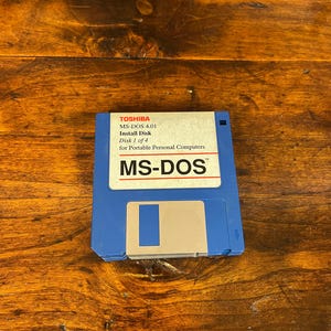 May include: A blue 3.5-inch floppy disk with a white label. The label reads TOSHIBA MS-DOS 4.01 Install Disk Disk 1 of 4 for Portable Personal Computers and features the text MS-DOS in a large bold font above a silver metal sliding shutter.