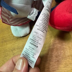 May include: A plush doll with a white face, red and blue outfit, and a white bonnet with lace trim. The doll has a tag with text in multiple languages, including English and Japanese. The doll's outfit has red and white stripes.