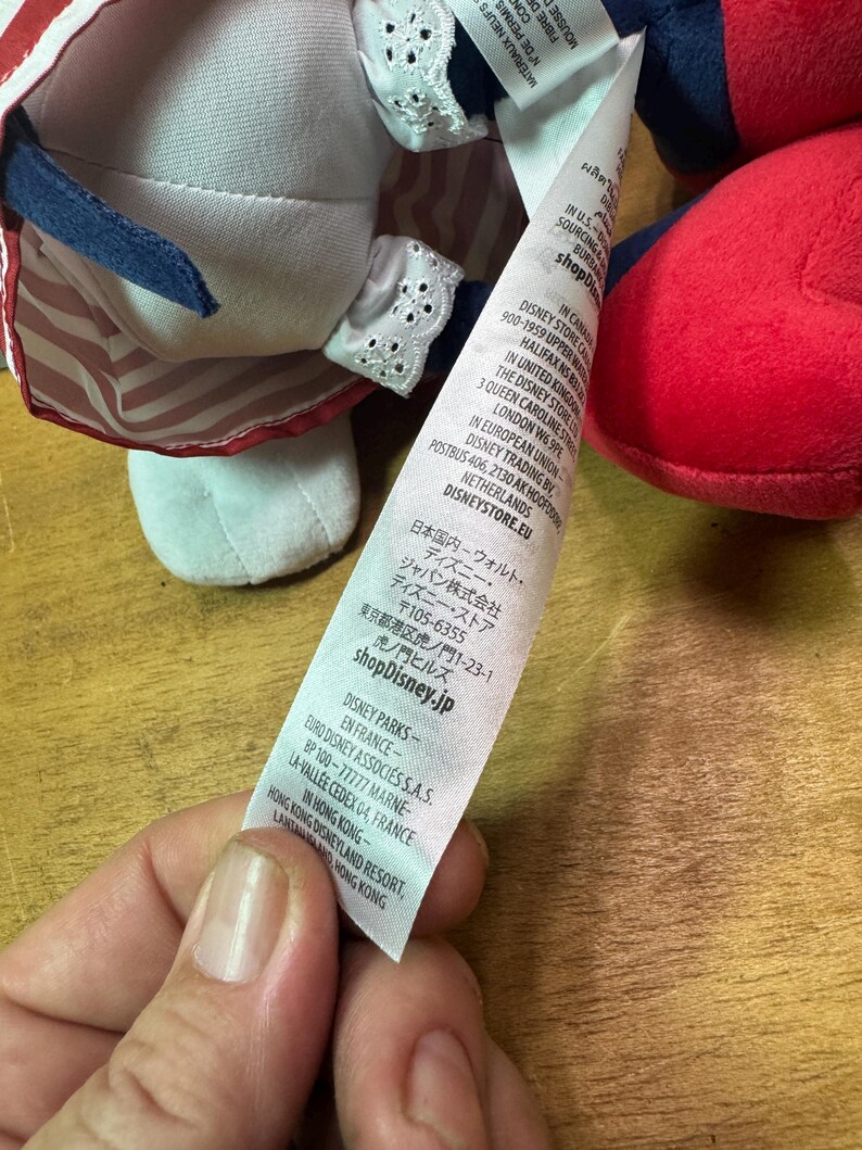 May include: A plush doll with a white face, red and blue outfit, and a white bonnet with lace trim. The doll has a tag with text in multiple languages, including English and Japanese. The doll's outfit has red and white stripes.