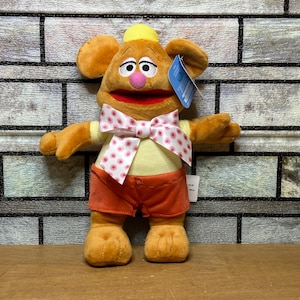 May include: A plush toy character with large ears, a yellow hat, and a pink nose. The character wears an orange outfit with a white bow tie decorated with pink polka dots. The toy is standing in front of a brick wall.