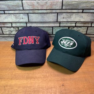 Op de afbeelding: Twee baseballpetten. Eén is marineblauw met "FDNY" in rode blokletters. De andere is donkergroen met een wit en groen logo. Beide petten hebben gebogen kleppen en zijn gemaakt van een stevige stof.