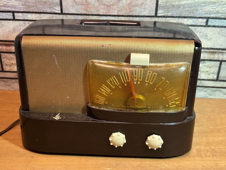 K&ouml;nnte beinhalten: Ein Vintage-Radio in Schwarz und Gold mit einem runden Zifferblatt und einem Drehknopf. Das Radio hat ein schwarzes Kunststoffgeh&auml;use und einen goldenen Metallgitter.