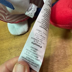May include: A close-up of a plush doll with a red and white striped dress and a blue bow. The doll's tag includes the text "shopDisney.jp" and other international Disney information.