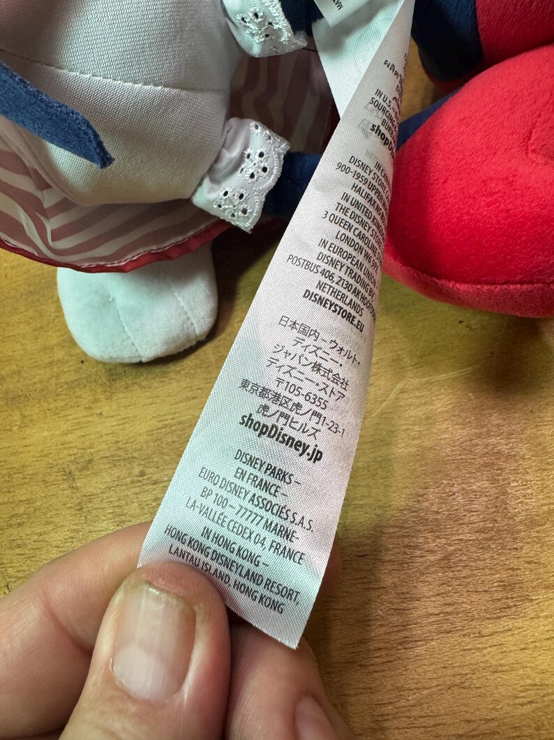 May include: A close-up of a plush doll with a red and white striped dress and a blue bow. The doll's tag includes the text "shopDisney.jp" and other international Disney information.