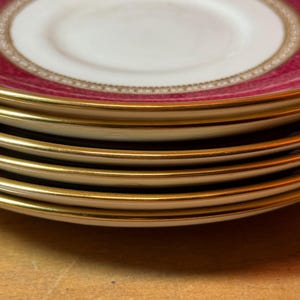 Puede incluir: Una pila de cuatro platillos blancos con ribete dorado. El platillo superior tiene un borde rosa con un patrón dorado y blanco.