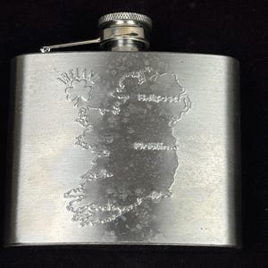 May include: A stainless steel hip flask featuring an etched map of Ireland, including Belfast, Dublin, and Cork. The word "Ireland" is above the map. The flask has a silver cap and is designed to hold liquids.