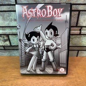 May include: A black and white DVD case for the Astro Boy The Classic 1960s Children's TV Series. The cover features illustrations of Astro Boy and a female character standing in a city, with the number 1 in a red circle at the bottom right.