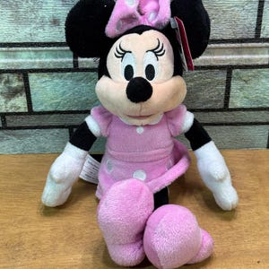 Puede incluir: Mu&ntilde;eca de peluche de Minnie Mouse sentada. La mu&ntilde;eca tiene orejas negras, un lazo rosa con lunares blancos y un vestido rosa con lunares blancos. Los brazos y las piernas de la mu&ntilde;eca son blancos y rosas. La mu&ntilde;eca est&aacute; sobre una superficie de madera.