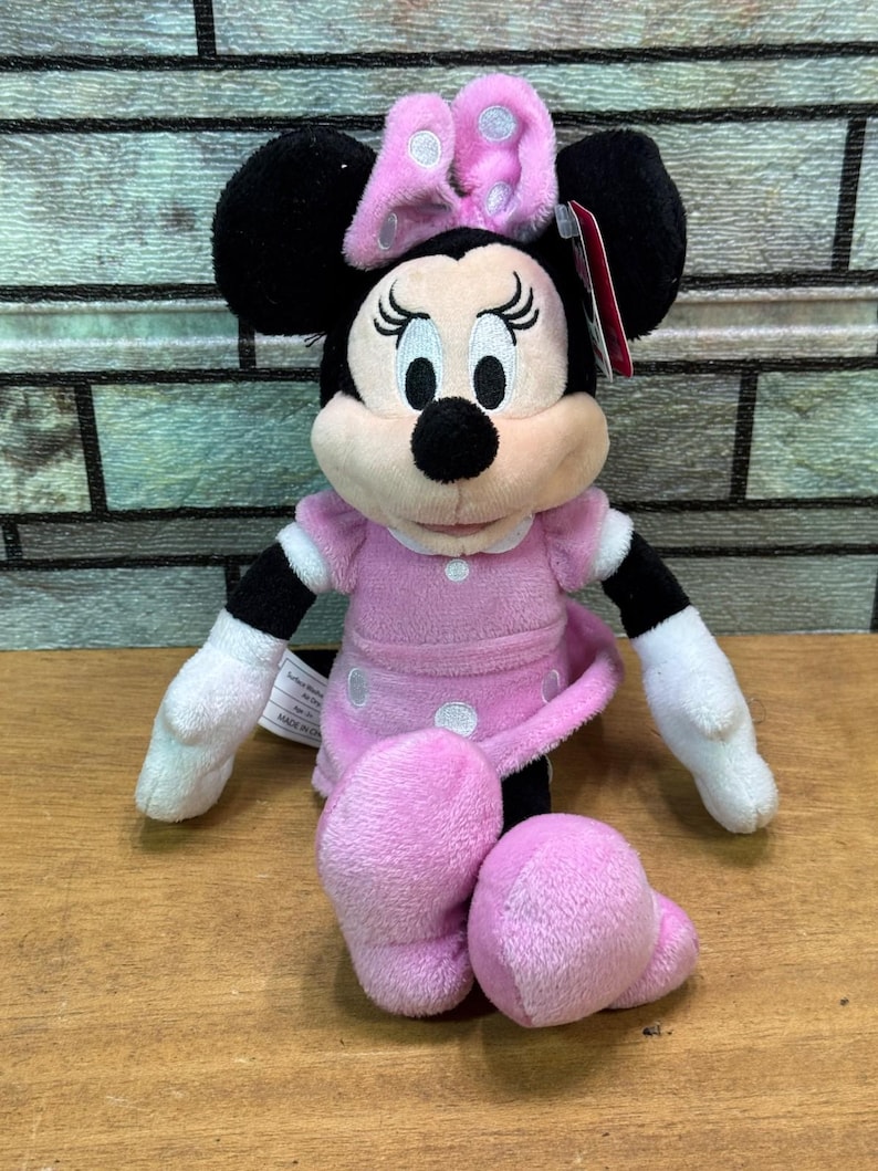 Puede incluir: Mu&ntilde;eca de peluche de Minnie Mouse sentada. La mu&ntilde;eca tiene orejas negras, un lazo rosa con lunares blancos y un vestido rosa con lunares blancos. Los brazos y las piernas de la mu&ntilde;eca son blancos y rosas. La mu&ntilde;eca est&aacute; sobre una superficie de madera.