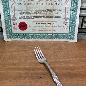 Puede incluir: Un tenedor de mesa plateado con un diseño de mango floral ornamentado, acompañado de un certificado de garantía de papel vintage de Wm. Rogers Mfg. Co. El certificado presenta bordes decorativos verdes y texto impreso en negro.