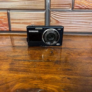 以下が含まれることがあります： フロントスクリーン、4.5-22.5mm 1:2.5-6.3 25mm仕様のSamsungレンズ、フラッシュ、Wi-Fiロゴを備えた黒いSamsungデジタルカメラ。このコンパクトなデバイスの前面には、Samsungのブランド名とHDのラベルが表示されています。