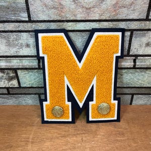 May include: A varsity-style letter M patch featuring a textured gold chenille center, a white felt border, and a dark navy blue felt backing. Two small, round, gold-colored metallic basketball-shaped buttons are attached to the bottom of the letter.